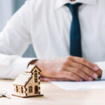 Osoba w białej koszuli i ciemnym krawacie siedzi przy biurku z modelem domu i kluczami, podpisując dokumenty związane z nieruchomościami