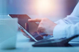 Dłonie korzystające z tabletu na tle cyfrowej grafiki, symbolizujące pracę z danymi i technologiami.