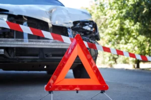 Uszkodzony samochód po kolizji drogowej z ustawionym trójkątem ostrzegawczym i taśmą zabezpieczającą miejsce wypadku.